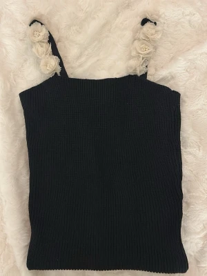 Svart stickad linnetopp från Zara - Säljer en svart stickad linnetopp från Zara med axelband dekorerade med stora, krämvita tygblommor. Toppen är ribbad och har en ärmlös design som ger en cool och trendig vibe. Perfekt för att lyfta din outfit med unika detaljer.