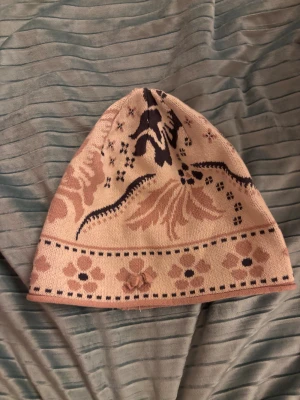 Blommig stickad mössa i beige/rosa och svart - Två stickade mössor med blommigt mönster och dekorativ rosett framtill. Ena mössan är beige med bruna och svarta detaljer, den andra är mörkgrå med svarta och beige inslag. Båda har en mjuk och skön passform, perfekta för att ge din look en unik touch.