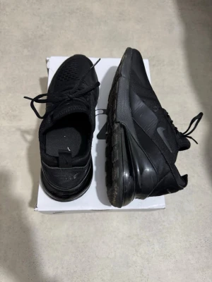 Nike Air Max 270 svarta sneakers - Nike Air Max 270 sneakers i helsvart färg med stickad ovandel och stor synlig Air-enhet i hälen. Snygg, sportig design med snörning och platt sula. Perfekt för dig som gillar streetstyle och vill ha bekväma skor med modern look.