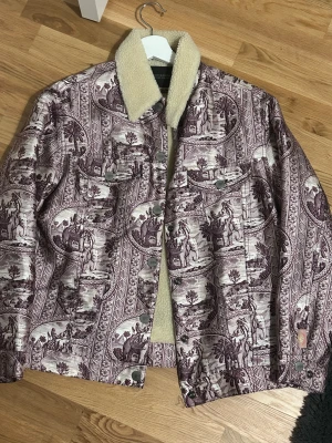 Mönstrad teddyjacka Scotch & Soda - Unik teddyjacka från Scotch & Soda med lila och beige vintageinspirerat mönster med elefanter och palmer. Jackan har fluffigt teddyfoder och krage, knappar framtill och två bröstfickor. Perfekt för dig som vill sticka ut med coola detaljer och skön värme.