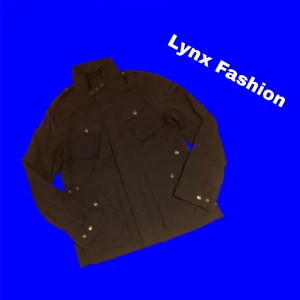 Svart vindjacka från Woolrich M - Svart vindjacka från Woolrich med hög krage, flera fickor med knappar och dragkedja framtill. Jackan har en stilren look och är tillverkad i lätt polyester, perfekt för blåsiga dagar. Insidan är fodrad i blått tyg för extra komfort.