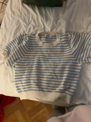 Randig stickad topp från A Place Apart - Säljer en söt stickad topp från A Place Apart med korta ärmar och rund hals. Toppen har ett randigt mönster i ljusblått och vitt samt ribbade kanter vid hals, ärmslut och nederkant. Perfekt för vår och sommar!