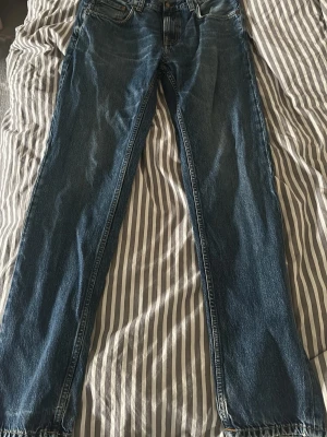 Blå gritty Jackson jeans från Nudie Jeans, 31/32 - Säljer ett par klassiska blå jeans från Nudie Jeans i storlek 31/32. Modellen har raka ben och fem fickor, med Nudie Jeans logga på bakfickan och läderpatch i midjan. Jeansen är tillverkade i slitstark bomull och har en snygg tvättad finish.