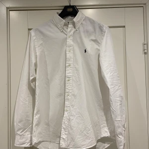 Vit skjorta från Ralph Lauren L - Vit skjorta från Ralph Lauren i storlek L, custom fit. Klassisk button-down krage och broderad logga på bröstet. Skjortan är långärmad och har knappar hela vägen framtill. Tillverkad i mjuk bomull som känns skön mot huden.