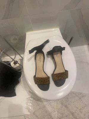 Svarta sandaler med leopardmönster - Säljer ett par stilrena sandaler från Clarks med svart sula och rem samt en bred rem över tårna i leopardmönstrad mocka..