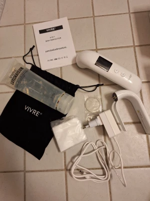 VIVRE 6 in 1 Skin Perfector - VIVRE 6 in 1 Skin Perfector med vit apparat, fuktgel, laddare, bomullspads, svart tygpåse och manual. Perfekt för ansiktsvård och återfuktning, med digital display och flera funktioner för hudvård. Gelén är transparent och kommer i en tub på 300g.