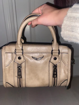 Beige handväska med dragkedjor - Säljer en beige handväska i skinnimitation med två rejäla handtag och snygga svarta detaljer. Väskan har dekorativa dragkedjor framtill och ett litet emblem i silver. Perfekt storlek för det viktigaste och har en cool, boxig form.