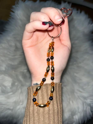 Hemgjord nyckelring🌞 - Hemgjord nyckelring med pärlor i guld, orange och brun/svart, samt ett hjärtformat fäste i metall. Perfekt smycke till väskan (bagcharm), nyckelring.