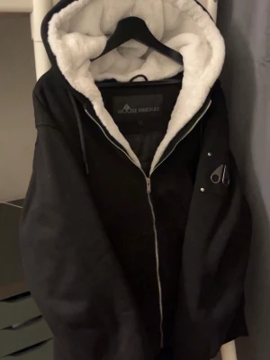 Svart Moose Knuckles Bunny  - Svart jacka/hoodie/cardigan från Moose Knuckles med vit fluffig huva och snyggt emblem på ärmen. Köpt från johnells om du vill se kvitto eller har några frågor säg till. Jackan har dragkedja framtill och mönstrat innerfoder i satin. Perfekt för dig som vill ha en varm och stilren look med coola detaljer. VÅGA LÄGG PRISFÖRLAG