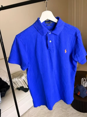 Blå pikétröja från Polo Ralph Lauren - Snygg blå pikétröja från Polo Ralph Lauren med klassisk krage och korta ärmar. Tröjan har två knappar framtill och den ikoniska broderade loggan på bröstet. Perfekt för en stilren och sportig look.