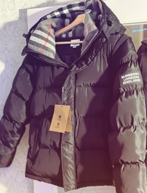 Svart pufferjacka från Burberry - Svart pufferjacka från Burberry med avtagbara ärmar och huva fodrad med klassiskt rutigt mönster i grått, vitt och rött. Jackan har dragkedja, knappar och två fickor med zip framtill. Perfekt för dig som vill ha en snygg och mångsidig jacka.🧥🔥