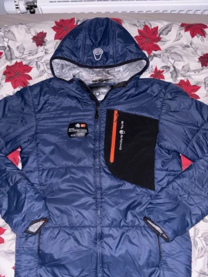 Blå Sail Racing pufferjacka med huva - Blå pufferjacka från Sail Racing med huva och coolt Sail Antarctica-märke på bröstet. Jackan har svart panel med orange dragkedja på vänster sida och två sidofickor med dragkedja. Materialet är polyester och jackan är perfekt för kalla dagar.