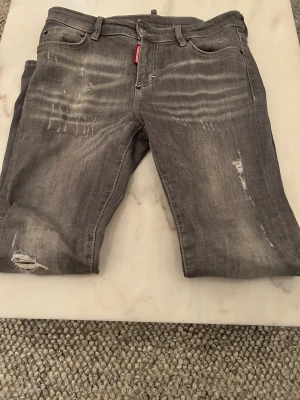 Grå jeans från Dsquared2, storlek 40 - Säljer ett par grå jeans från Dsquared2 med slitna detaljer och snygga slitningar framtill och baktill. Klassisk femficksmodell med Dsquared2-logga på bakfickan. Jeansen har normal passform och är tillverkade i bomullsmaterial för en skön känsla. Hör av er vid minsta lilla fundering och mer bilder kan du också få !