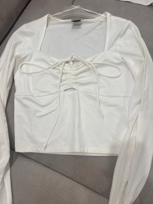 Vit långärmad topp med snörning Gina Tricot - Supersnygg vit långärmad topp från Gina Tricot med snörning framtill och liten cut-out under bysten. Toppen har slitsar vid ärmsluten och är gjord i mjukt bomullsmaterial. Perfekt att styla med jeans eller kjol för en trendig look.