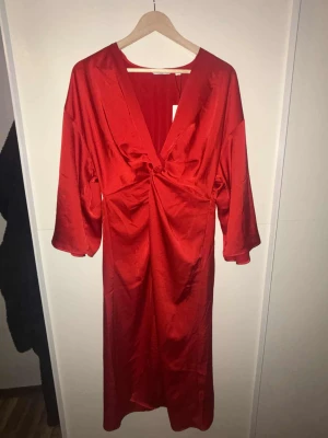 Röd långklänning med v-ringning - Snygg röd långklänning med djup v-ringning och trekvartsärmar. Klänningen har ett elegant fall och ett twistat parti framtill som ger en cool detalj. Materialet har en lätt glansig finish som känns lyxig och passar perfekt när du vill sticka ut. Inte använd