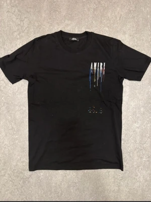 Svart AMIRI t-shirt med färgstänk - Svart t-shirt från AMIRI med stort tryck på ryggen och mindre tryck på bröstet. Trycket består av AMIRI-loggan med färgstänk i blått, vitt, rött och gult som rinner ner. Rund hals och korta ärmar. Materialet är mjuk bomull, perfekt för en cool streetwear-look.