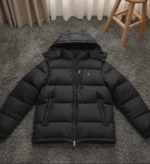 Svart pufferjacka från Polo Ralph Lauren - Svart pufferjacka från Polo Ralph Lauren med röd broderad logga på bröstet. Jackan har huva med snörning, dragkedja och knappar framtill samt två sidofickor med dragkedja. Materialet är syntet och jackan har en quiltad look som ger extra volym. Kvitto finns