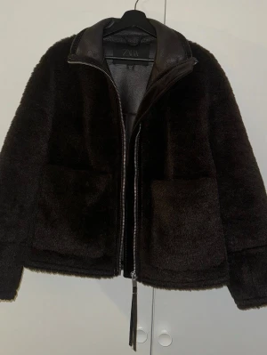 Svart teddyjacka från Zara - Mjuk och fluffig svart teddyjacka från Zara med detaljer i skinnimitation vid kragen och dragkedja framtill. Jackan har två stora fickor och en stilren look som passar perfekt när du vill vara både varm och snygg. Perfekt för kyliga dagar.