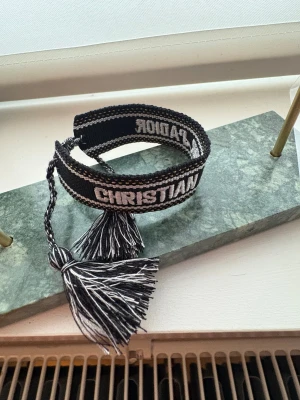 Christian Dior svart tygarmband - Snyggt svart armband från Christian Dior med broderad text 'J'ADIOR' och 'CHRISTIAN DIOR' i vitt. Armbandet är gjort i tyg med flätade snören och två stora tofsar i svart och vitt. Perfekt statement piece med cool design och exklusiv känsla.