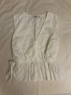 Vit omlottopp med broderade blommor NA-KD - Superfin vit omlottopp från NA-KD med broderade blommor och volangkant nedtill. Toppen är ärmlös och har knyt i sidan för en snygg passform. Perfekt för sommarens alla tillfällen, lätt och luftig i bomull.