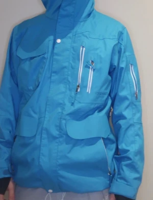 Blå skidjacka med huva från Warp - Blå vindjacka från Warp med huva och flera praktiska fickor med dragkedja. Jackan har lång ärm, vita detaljer på dragkedjorna och justerbar huva. Perfekt för dig som gillar att åka skidor och vill ha en snygg och funktionell jacka.