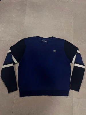 Blå sweatshirt från Lacoste Sport - Säljer en snygg marinblå sweatshirt från Lacoste Sport med klassisk krokodillogga på bröstet. Tröjan har långärmade svarta ärmar med vita och blå ränder som ger en sportig vibe. Rund halsringning och mjukt bomullsmaterial. Perfekt för chill dagar.