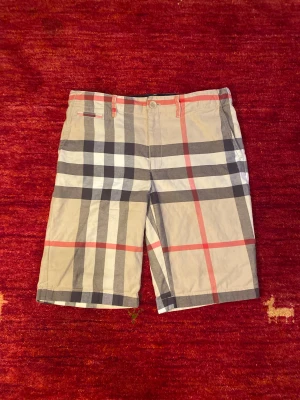 Burberry Shorts - Eleganta och tidlösa Burberry-shorts i klassiskt beige med det ikoniska rutmönstret i grått, vitt och rött. En stilren design som är direkt igenkännbar och aldrig går ur tiden. Tillverkade i ett lätt och bekvämt material, perfekta för vår- och sommarsäsongen.