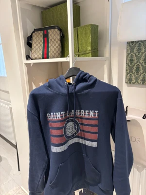 Mörkblå Saint Laurent hoodie - Snygg mörkblå hoodie från Saint Laurent med tryck i rött och vitt framtill. Tröjan har huva med snörning, stor magficka och långa ärmar. Perfekt för dig som gillar streetwear och vill ha en exklusiv vibe i garderoben.
