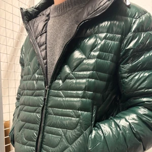 Mörkgrön quiltad dunjacka - Snygg mörkgrön dunjacka med quiltad design och glansig yta. Jackan har dragkedja framtill och är tillverkad i ultralätt polyamid med fyllning av 90% ankdun och 10% fjäder. 