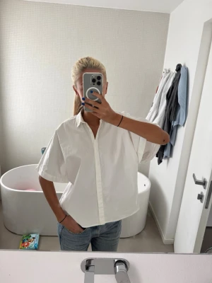 Vit loose skjorta med kort ärm - Säljer en vit skjorta med klassisk krage och korta ärmar. Skjortan har en loose passform och knäpps med knappar framtill. Materialet känns som bomull och är lätt och luftigt, perfekt för varmare dagar. Enkel och stilren design som är lätt att matcha.