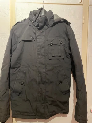 Svart parkajacka från Jack & Jones - Svart parkajacka från Jack & Jones med hög krage och flera fickor med knappstängning. Jackan har långa ärmar och en cool, urban look. Perfekt för dig som gillar streetstyle och vill ha en jacka med snygga detaljer och praktiska fickor.