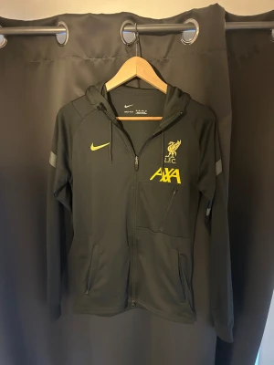 Svart Nike Liverpool hoodie XS - Svart hoodie från Nike med Liverpool FC-logga och gul AXA-tryck på bröstet. Dri-FIT-material som andas, hel dragkedja, huva med snörning och två fickor med dragkedja. Snygga gula detaljer och broderad Nike Swoosh på framsidan.