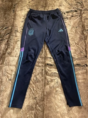 Adidas Argentina träningsbyxor XS - Mörkblå träningsbyxor från Adidas med Argentina-logga och tre ljusblå ränder längs benen. Byxorna har lila detaljer vid knäna, elastisk midja och smal passform. Perfekta för fotboll eller träning, tillverkade i polyester.