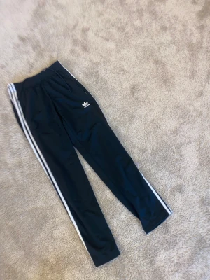 Svarta Adidas Primeblue byxor XS - Svarta Adidas byxor i modellen Primeblue med klassiska vita ränder längs sidorna och broderad logga framtill. Byxorna har resår i midjan och dragkedjor vid fickorna. Tillverkade av återvunnet material.