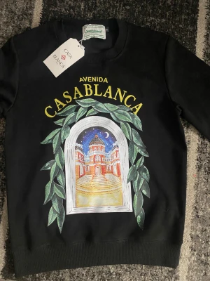 Svart sweatshirt från Casablanca - Säljer en svart sweatshirt från Casablanca med färgglatt tryck framtill som visar en byggnad omgiven av gröna blad och texten 'Avenida Casablanca' i gult. Tröjan har rund hals och långa ärmar. Perfekt för dig som gillar streetwear med unik design.