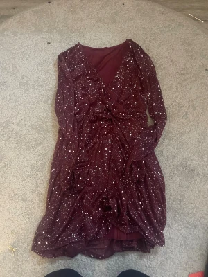 Vinröd glittrig omlottklänning - Supersnygg vinröd kortklänning med glittriga silverprickar och omlott-design. Klänningen har långärmade transparenta ärmar och ett mjukt innerfoder. Materialet är lätt och skimrande, perfekt för att sticka ut. V-ringad och med markerad midja för en trendig look.