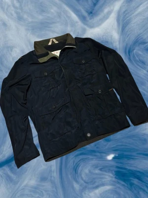 Svart vindjacka från Esprit S - Svart jacka från Esprit med fyra stora fickor framtill och knappar. Jackan har en krage i kontrasterande olivgrön färg. Perfekt för dig som gillar en clean och funktionell stil. Storlek S