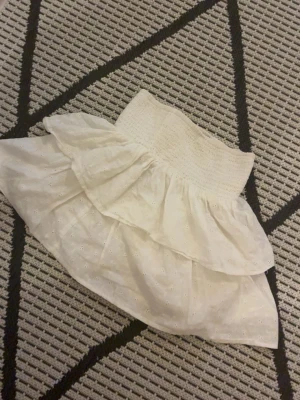 Vit kortkjol med volanger xxs - Supersöt vit kortkjol med bred smock i midjan och dubbla volanger. I storlek Xss.Kjolen sitter tajt vid midjan och är inte genomskinlig 