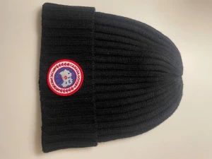 Svart ribbad mössa från Canada Goose - Säljer en svart ribbstickad mössa från Canada Goose med klassisk rund logotyp framtill. Mössan har bred uppvikt kant och är tillverkad i mjukt stickat material som håller dig varm under kalla dagar.