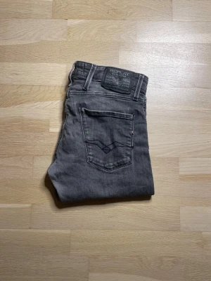 Replay Anbass grå jeans, storlek 30 - Snygga Replay Anbass jeans i en mörkgrå tvätt med klassisk femficksdesign och tydliga Replay-detaljer på fickor och patch. Jeansen har en slim passform och är tillverkade i mjukt jeanstyg med lite stretch för extra komfort. Den ända defekt som finns är att de är fixade hål som visas på bild 5.