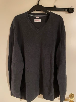 Mörkgrå v-ringad tröja från s.Oliver - Säljer en stilren mörkgrå v-ringad tröja från s.Oliver i storlek XXL. Tröjan har långa ärmar, diskret broderad logga på bröstet och ribbade muddar vid hals, ärmslut och nederkant. Perfekt för dig som gillar enkel och klassisk stil.