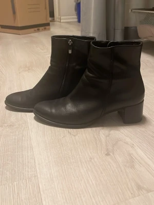 Svarta boots med klack och dragkedja - Äkta Ecco boots, Snygga svarta boots i skinn med rund tå och låg klack. De har dragkedja på sidan för enkel på- och avtagning. Stilren design som passar till många olika outfits och ger en cool vibe. Perfekt för dig som gillar klassiska och tidlösa skor. Har en liten skadda på klacken se bild 6 men påverkar inte funktionen av skorna ska tvättas innan jag skickar till köparen original pris 1700 sänkt pris till 1450 med tanke på skadan