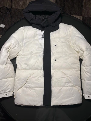 Vit Moncler pufferjacka med svart huva - Säljer en clean pufferjacka från Moncler i vit med svart huva och insida. Jackan har stora fickor, dragkedja och tryckknappar framtill. Moncler-logga broderad på huvan. Perfekt för dig som vill ha en stilren vinterlook.