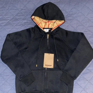 Svart hoodie med dragkedja från Burberry - Säljer en stilren svart hoodie från Burberry med dragkedja och justerbar huva. Insidan av huvan har det klassiska Burberry-mönstret i beige och rött. Perfekt för en avslappnad och trendig look. Står XL men sitter mer som M(A-cop)