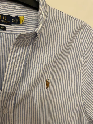 Randig skjorta från Polo Ralph Lauren - Snygg skjorta från Polo Ralph Lauren i klassisk passform, med ljusblå och vita ränder. Broderad logga på bröstet och knappar framtill. Tillverkad i mjuk bomull och har långärmade ärmar med manschett. Perfekt för dig som gillar stilrena och tidlösa plagg. Nypris ≈ 1500kr-1990kr