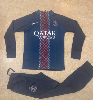PSG träningsset från Nike, marinblå - Säljer ett fett PSG träningsset från Nike i marinblått med långärmad tröja och matchande byxor. Tröjan har rött och vitt mönster framtill, PSG-logga och Qatar Airways-tryck. Byxorna har PSG-emblem och resår i midjan. Perfekt för fotbollsträning eller chill.