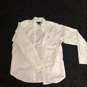 Vit skjorta från Ralph Lauren - Säljer en klassisk vit skjorta från Ralph Lauren med lång ärm och knappar framtill. Skjortan har den ikoniska broderade loggan på bröstet och är gjord i mjuk bomull. Perfekt för dig som gillar stilrena och tidlösa plagg.