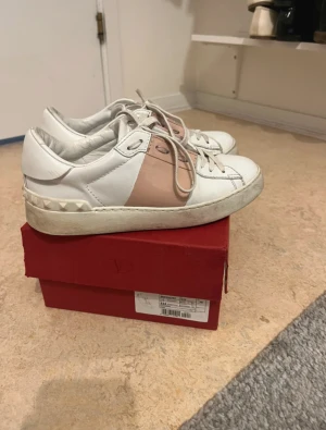 Valentino sneakers vit/ljusrosa - Snygga Valentino sneakers i vitt skinn med ljusrosa panel och klassiska nitar vid hälen. Rund tå, platt sula och snörning framtill. Perfekt för dig som gillar exklusiva märken och vill ha en unik detalj i din outfit.