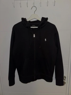 POLO RALPH LAUREN – Svart zip-hoodie i storlek S - Säljer en stilren och välsittande svart hoodie från Polo Ralph Lauren, i storlek S. Den har en praktisk dragkedja fram, mjuk och bekväm kvalitet, och den ikoniska Polo Ralph Lauren-loggan. En perfekt vardags- eller fritidsplagg som passar till det mesta.  Hoodien är i mycket gott skick, men eftersom den har blivit för liten för mig får den ett nytt hem. Ingen skada eller fläck.  Bor i Strängnäs. Produkten finns redo för upphämtning. Kan även skickas som ett brev eller paket inom Sverige (fraktko