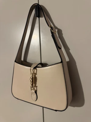 Beige axelväska med gulddetalj - Snygg beige axelväska Väskan har en justerbar axelrem och ett snyggt guldfärgat spänne framtill som ger en lyxig känsla. Perfekt för dig som gillar minimalistisk design och vill ha en väska som sticker ut. Helt ny!